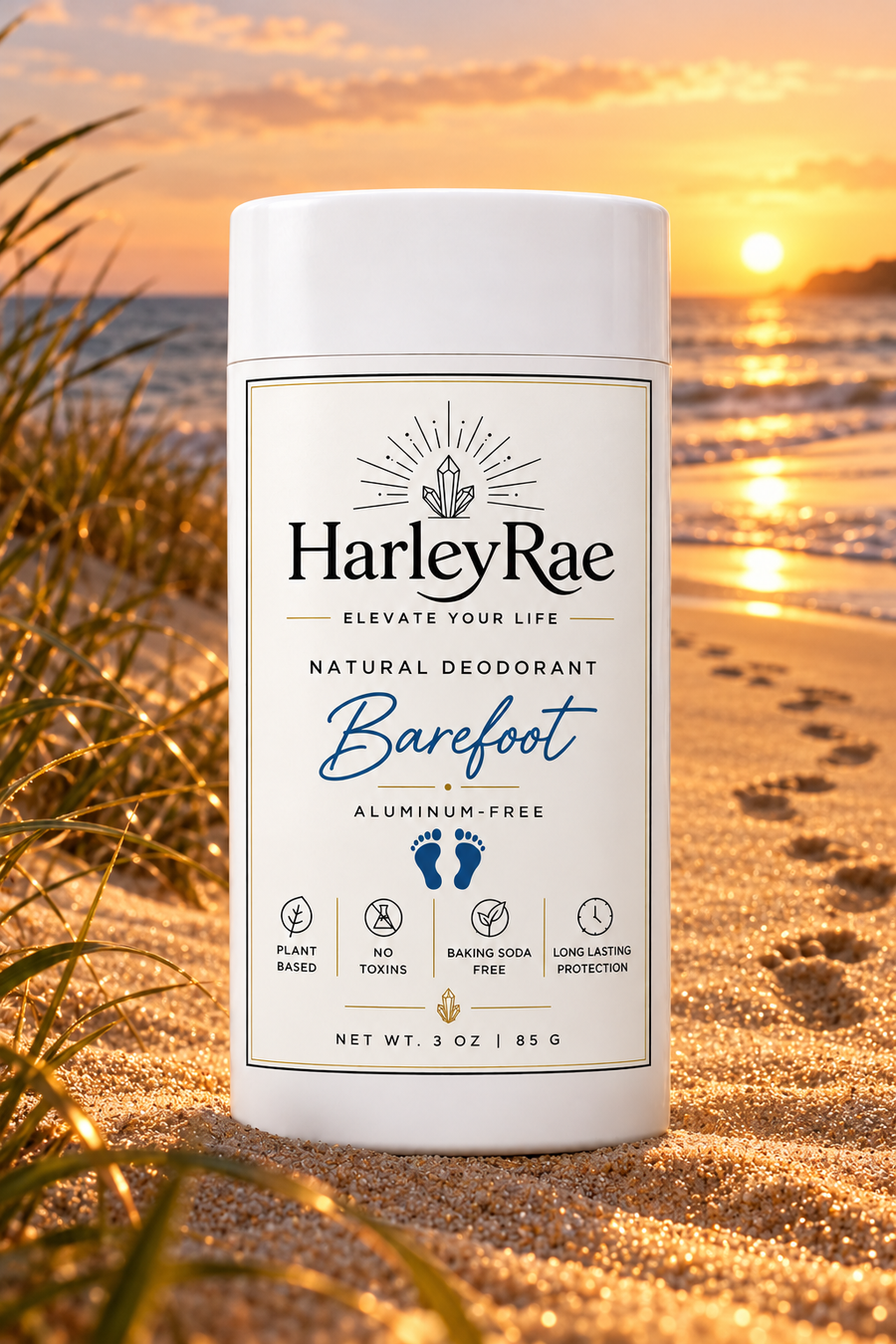 A white cylindrical bottle of HarleyRae Barefoot Natural Deodorant standing on a sandy beach at sunset, with ocean waves and grass blurred in the background.